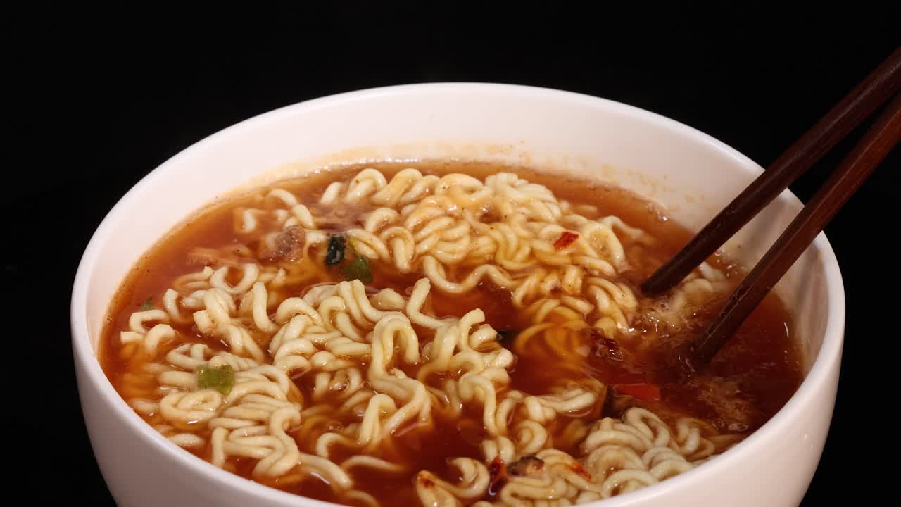 A pair of chopsticks lifts cooked instant noodles from a white bowl filled with broth, under even studio lighting and a black background