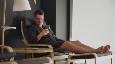 adult wealthy man is resting in spa salon or health center sitting in lounger and using smartphone viewing positive news