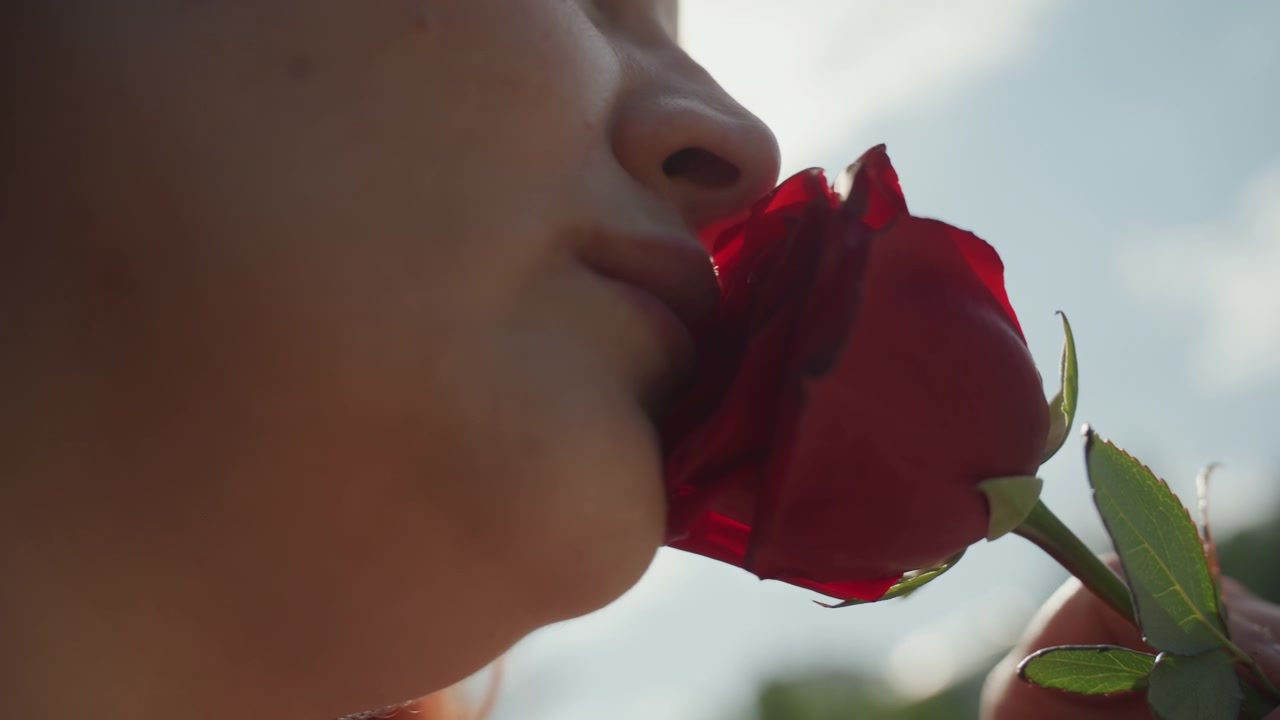 Primo piano di una donna che annusa e tiene in mano una rosa rossa all'aperto con la luce del sole. La mano afferra lo stelo mentre il viso si avvicina al fiore, catturando l'interazione tra il soggetto e il fiore. Lo sfondo mostra.