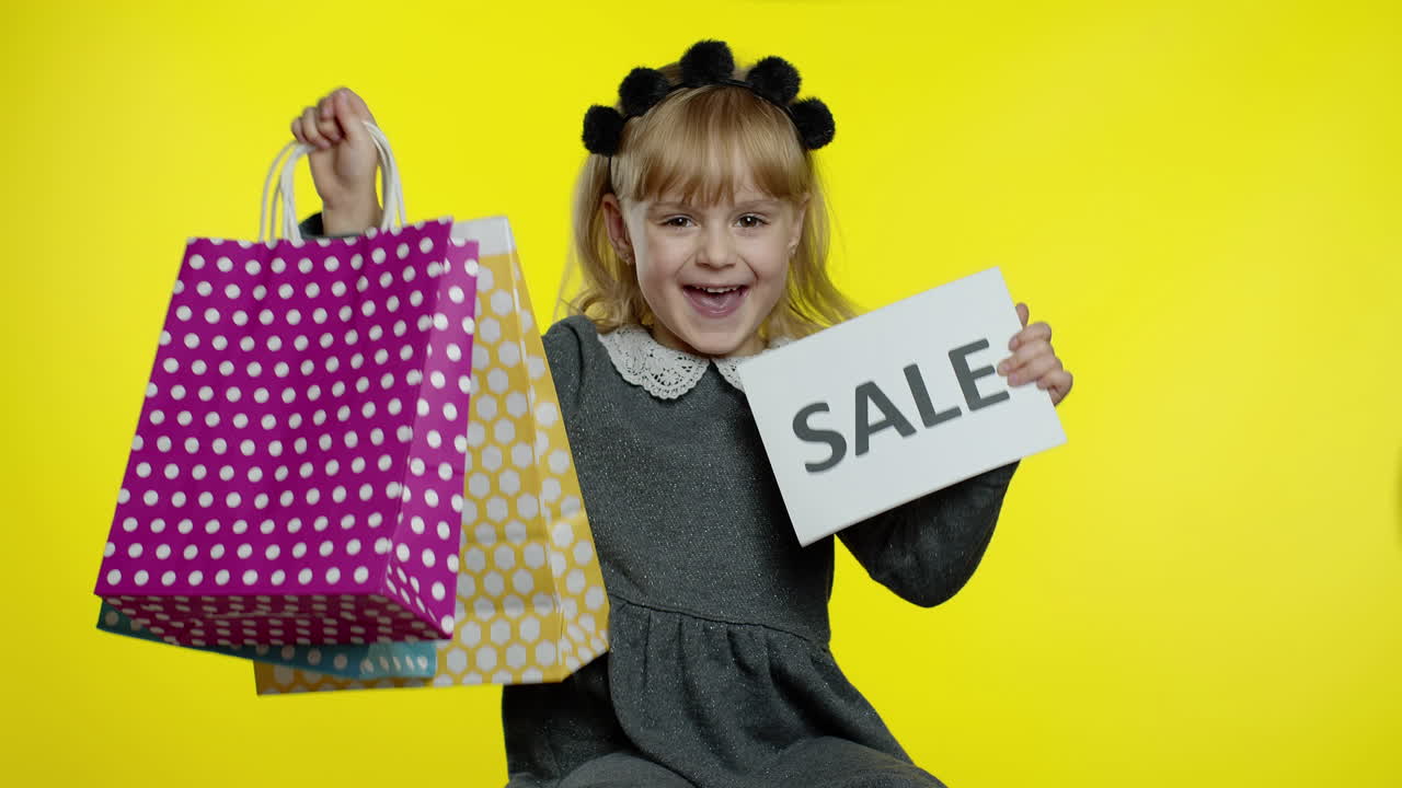 Child girl with shopping bags showing Sale word inscription note, smiling, rejoicing good discounts