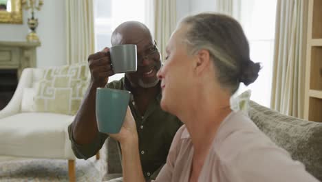 Happy-senior-diverse-couple-wearing-shirts-and-drinking-coffee-in-living-room