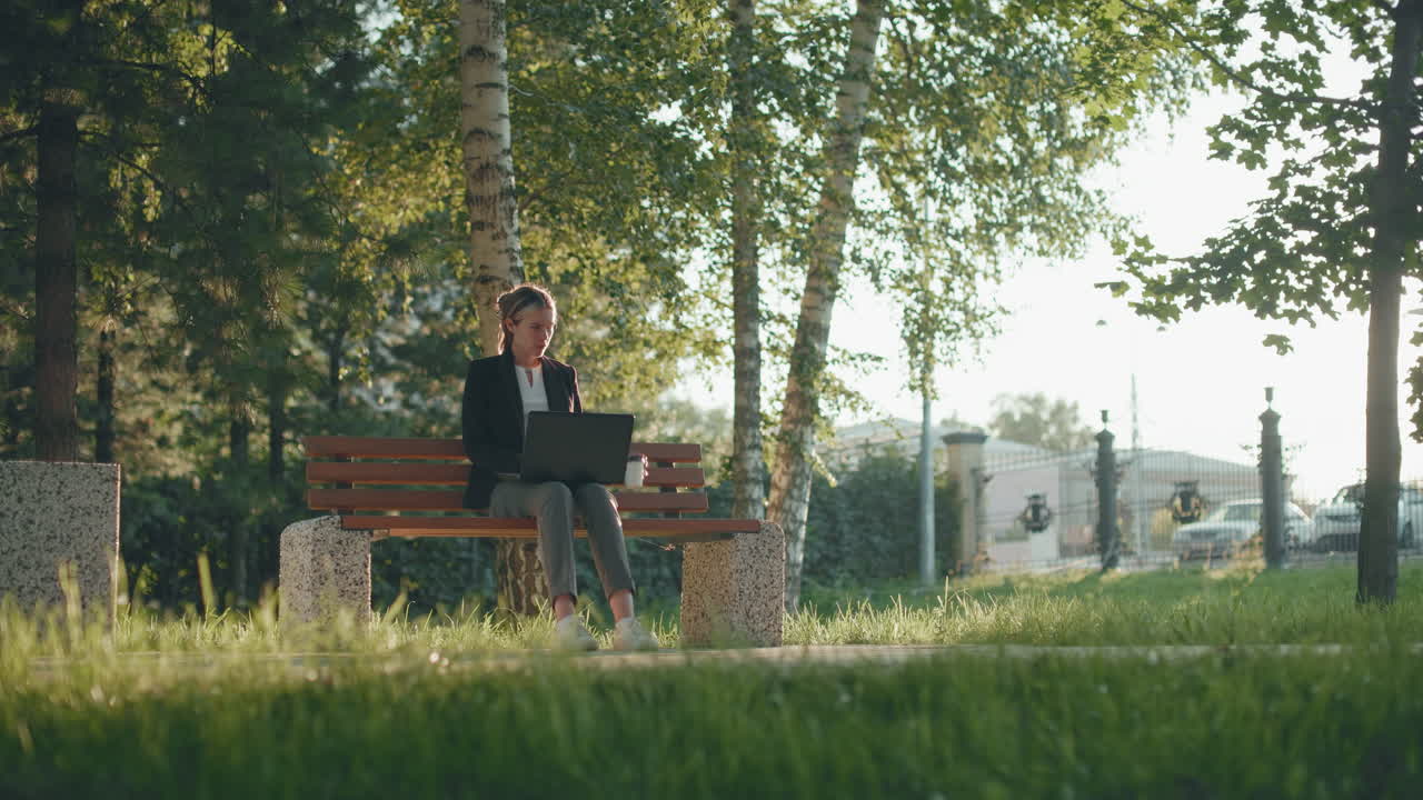 Freelancer seated outdoors on wooden bench working on system and sipping coffee with green trees, morning sunlight, and distant bus in urban park setting