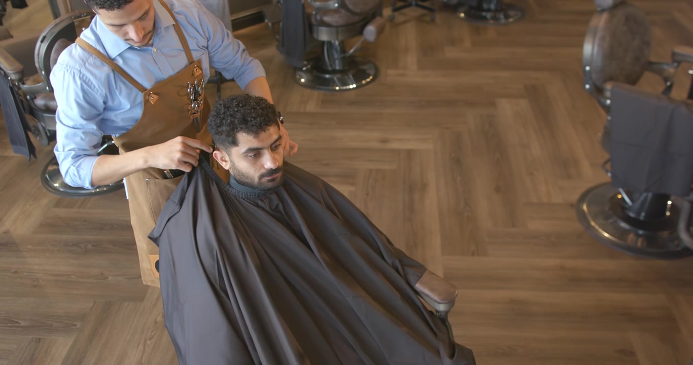 Mandlig barber trækker sort kappe over mandlig kunde i læderforklæde, forbereder klipning i barbersalon