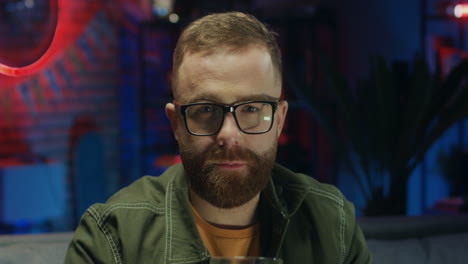 Portrait-Of-The-Young-Cheerful-Man-In-Glasses-Drinking-Beer-And-Doing-Cheers-Gesture-To-The-Camera-And-Smiling-In-The-Dark-Room