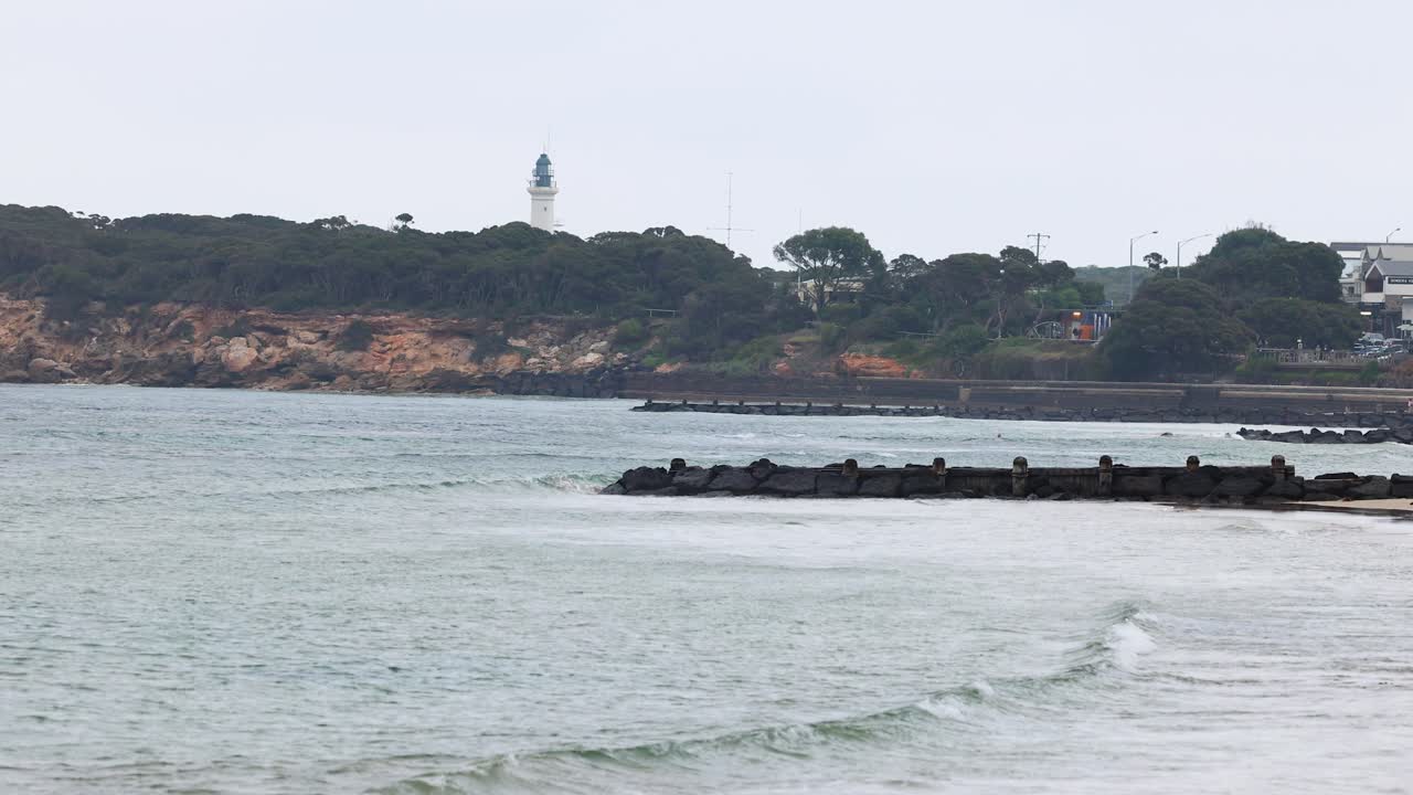 Calm ocean waves roll towards the Point Lonsdale lighthouse under overcast skies, creating a tranquil coastal scene
