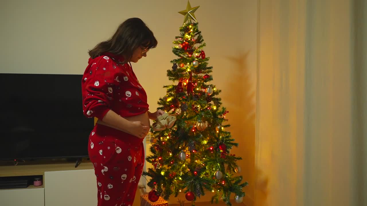 Linda cena de Natal de uma mulher vestindo roupas vermelhas com a barriga descoberta olhando para um par de sapatinhos de bebê em frente a uma árvore iluminada