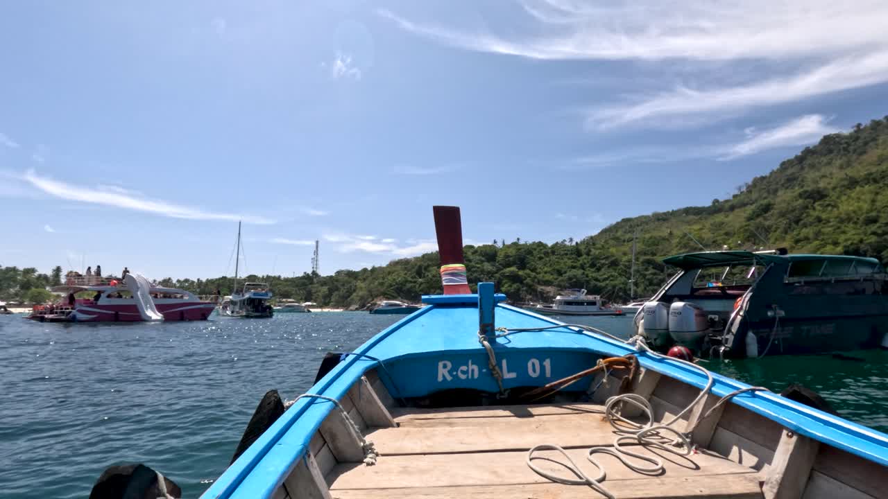 Wooden long-tail boat moves toward harbor, passing yachts under bright daylight and clear blue sky