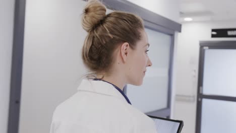 happy caucasian female doctor walking in hospital corridor using tablet, in slow motion