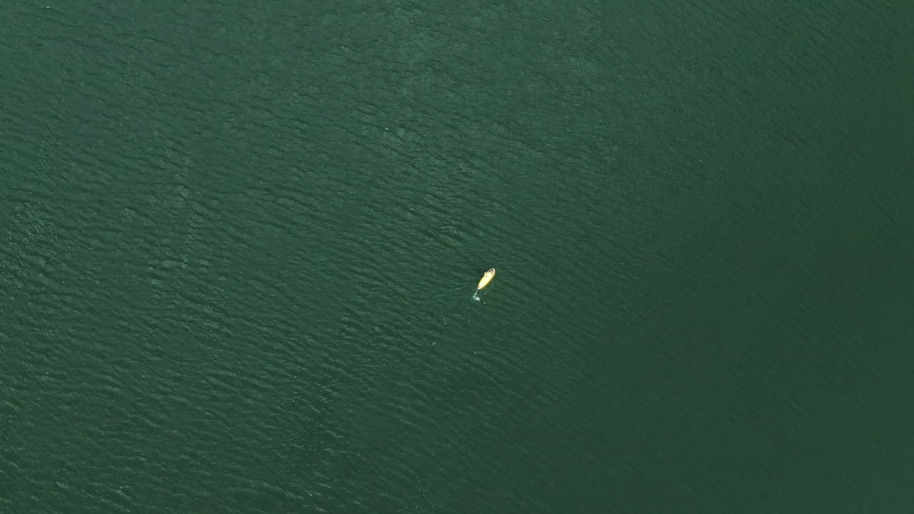persona remando en una tabla amarilla en el vasto agua del lago, vista aérea ascendente de arriba hacia abajo