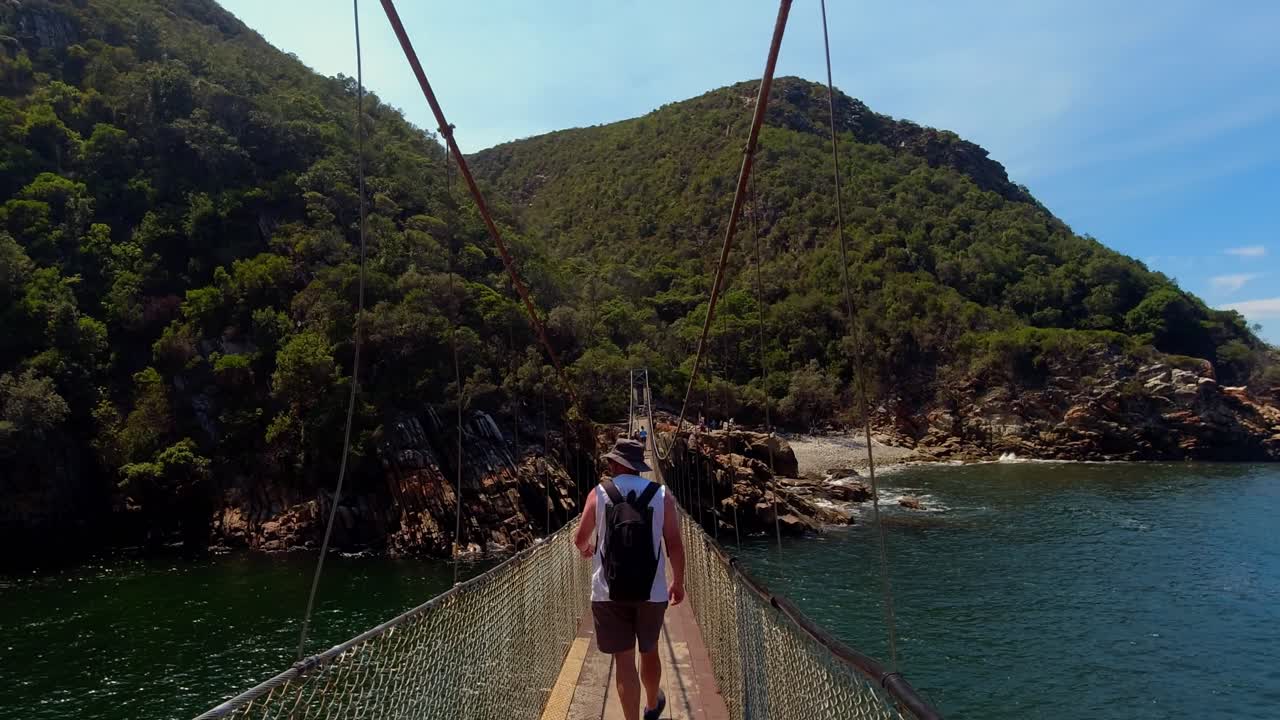 Um plano seguinte de um(a) mochileiro(a) atravessando a famosa ponte suspensa no Parque Nacional de Tsitsikamma, África do Sul. 4K
