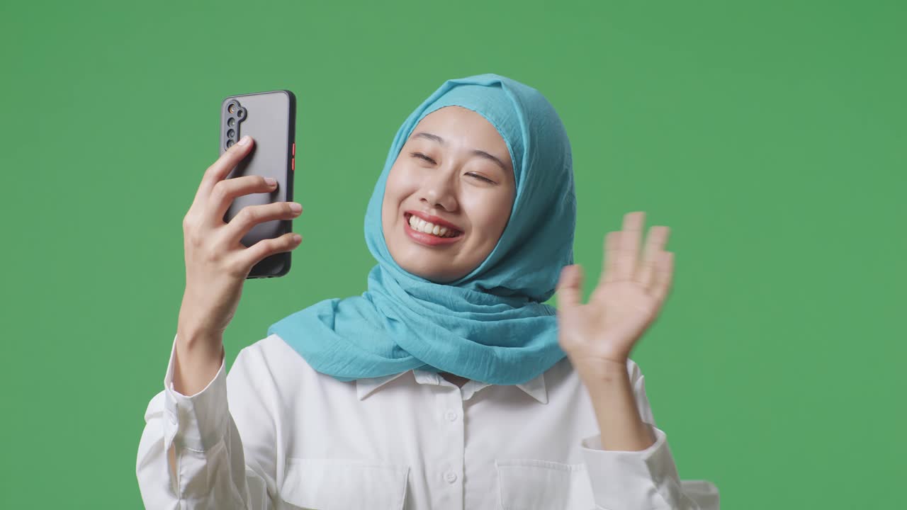 미소 짓는 아시아 무슬림 여성이 손을 흔들고 녹색 화면 배경 스튜디오에 서서 스마트폰으로 비디오 통화를 하고 있습니다.