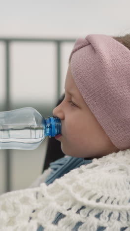 thirsty little girl drinks water from bottle sitting in wheelchair near mom on observation deck. woman enjoys walk with daughter with spinal injury closeup