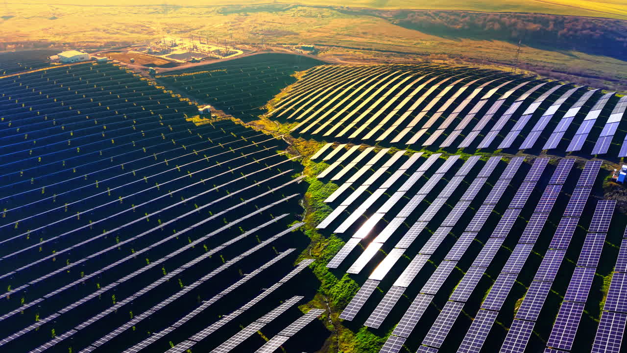 Fugleperspektiv af et stort solcelleanlæg på bakket terræn under den gyldne time, hvor solen glimter fra panelets overflader, og en transformerstation ses i baggrunden. Solenergipark ved solnedgang.