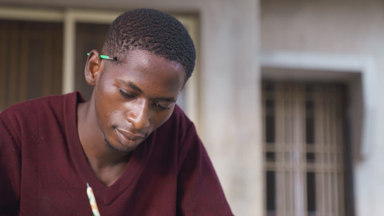 Uomo nero che dipinge con la matita, primo piano focalizzato di giovane artista africano che lavora su carta, matita dietro l'orecchio, luce soffusa, espressione contemplativa, carta testurizzata, studio esterno minimalista