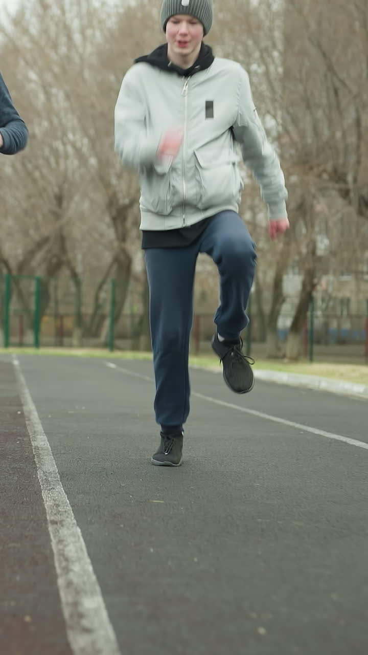 A coach guides a young boy through jogging exercises on a stadium track, by instructing him to lift his legs higher with bare trees and bar fence in the background