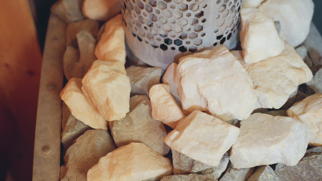 Close up of mixed sauna stones, pale and dark, arranged around perforated metal heater in steam room, capturing raw rocky textures, soothing earth tones
