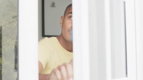 hombre biracial feliz bebiendo café y abriendo la ventana en una cocina brillante, cámara lenta