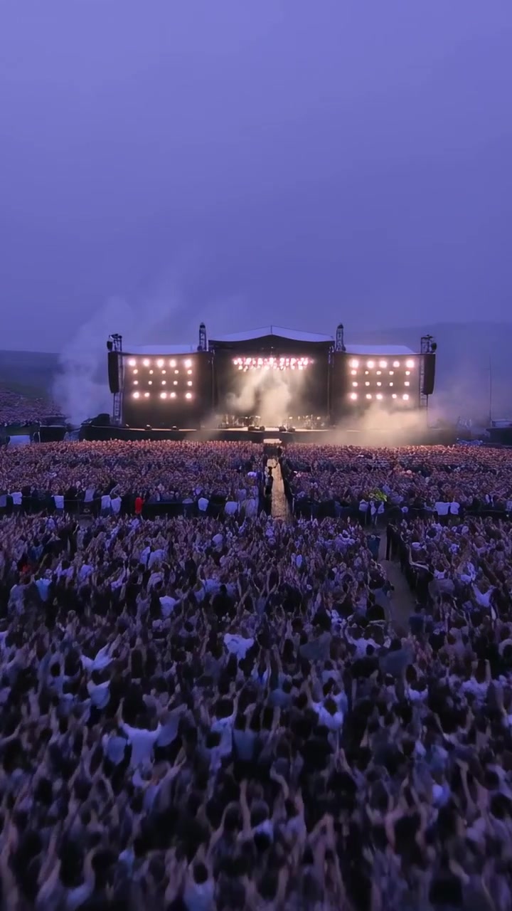 Video vertical: Multitud reaccionando con abrigos, levantando brazos y teléfonos en un campo al ritmo de la música, con luces ascendiendo.
