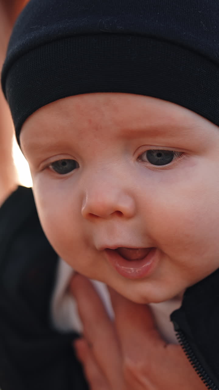 Face of an adorable Caucasian baby with grey eyes. Kid in black cap in mom's hands outdoors. Close up. Vertical video