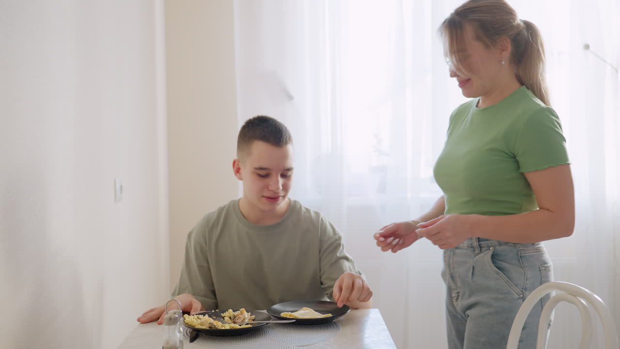 Caring mother in green shirt and jeans carries egg on black plate in bright kitchen, preparing to serve boy seated at dining table, natural daylight through curtain