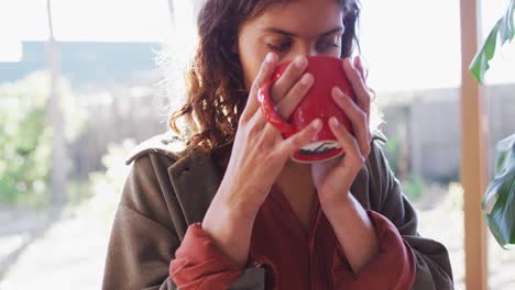 Thoughtful-mixed-race-woman-sitting-drinking-tea-in-sunny-cottage-kitchen