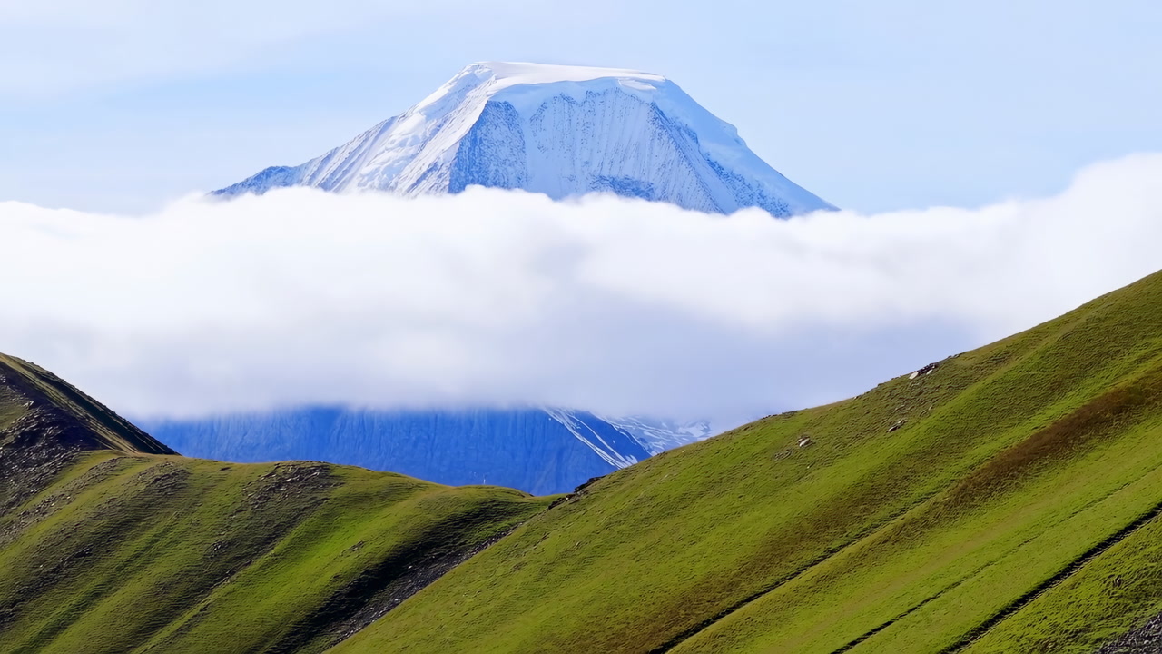 Snow Capped Mountain Above Low Clouds, Rolling Green Hills In Foreground, Soft Morning Light Accentuating