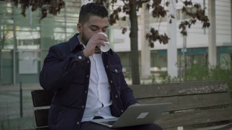 hombre bebiendo café para ir y usando computadora portátil al aire libre