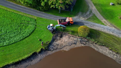 Aerial over excavator machines work next to a lake, construction outskirts, landscape of green fields