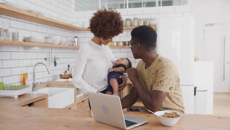 busy family in kitchen at breakfast with father working on laptop and mother caring for baby son