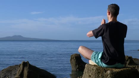 Un-Joven-En-Forma-Meditando-En-Una-Pose-De-Yoga-Sobre-Rocas-En-La-Playa-Por-La-Mañana