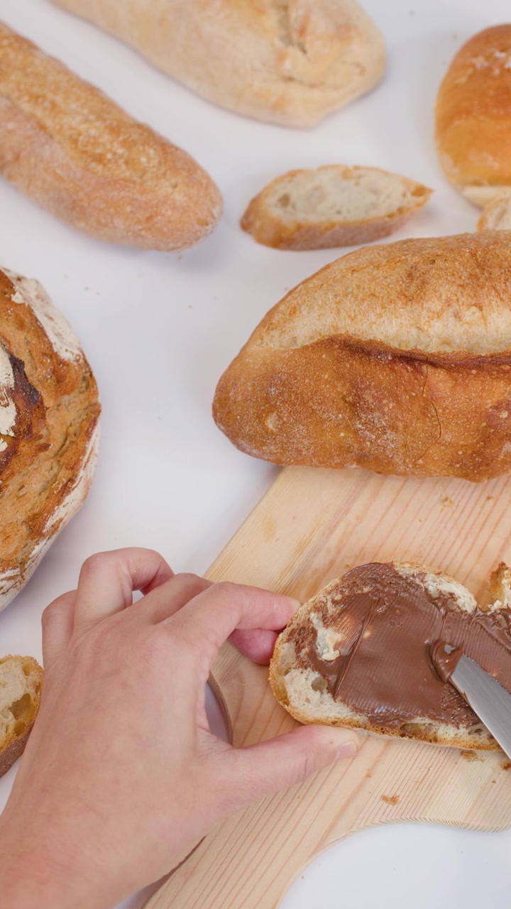 Close-up of hand spreading chocolate cream on bread over white background with assorted loaves.