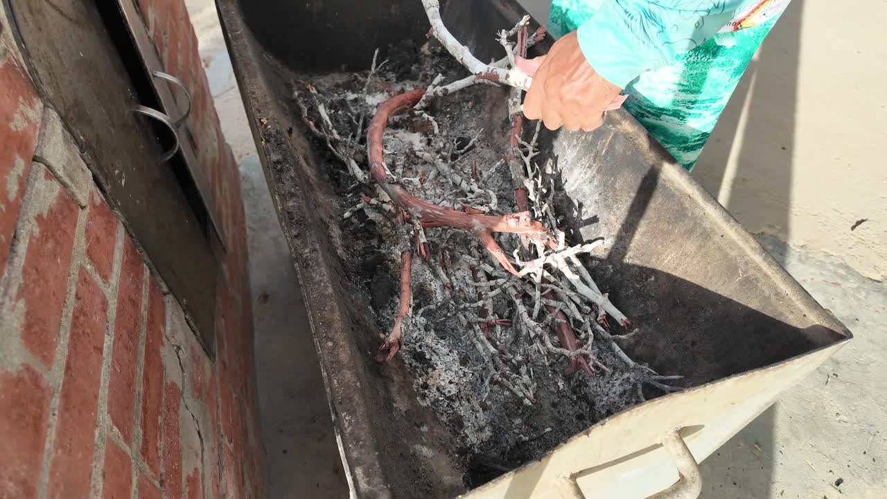 High-angle, textured detail of dry wood and small branches nestled into a bed of white ash in a metal container, ready for lighting the perfect charcoal fire