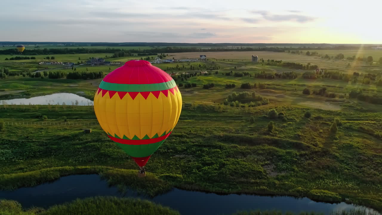 Travel in the air at sunset. Hot air balloon flying in the countryside. Colorful aerostat over the fields and lakes. Aerial view. Motion camera around.