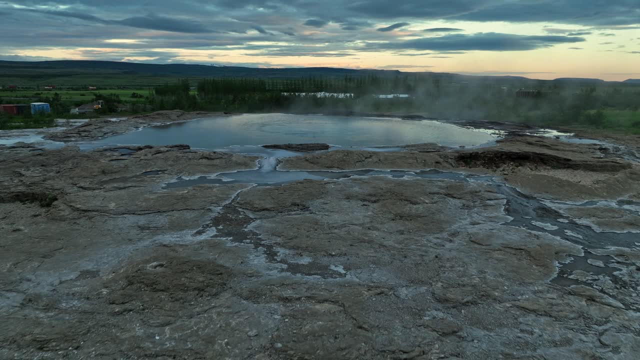 fotografía de avión no tripulado de las aguas termales de geysir en islandia con vapor que se eleva al atardecer, rodeado de terreno geotérmico y vegetación distante.
