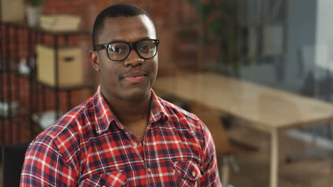 Portrait-Of-The-Young-Handsome-Man-In-Glasses-And-Red-Motley-Shirt-Sitting-In-Front-Of-The-Camera-And-Smiling