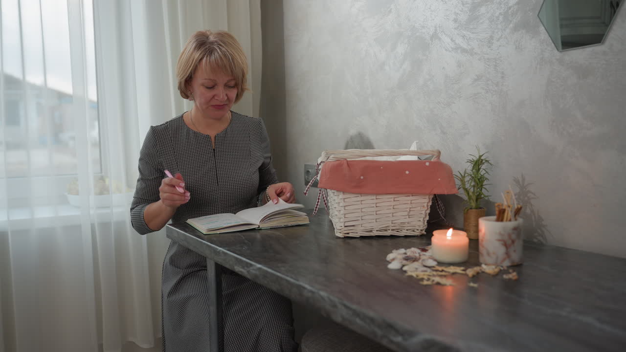 Young adult seated at table adjusting while opening book in hand holding pen next to basket candle seashells and plant in bright cozy room with soft curtains natural light and textured wall decor