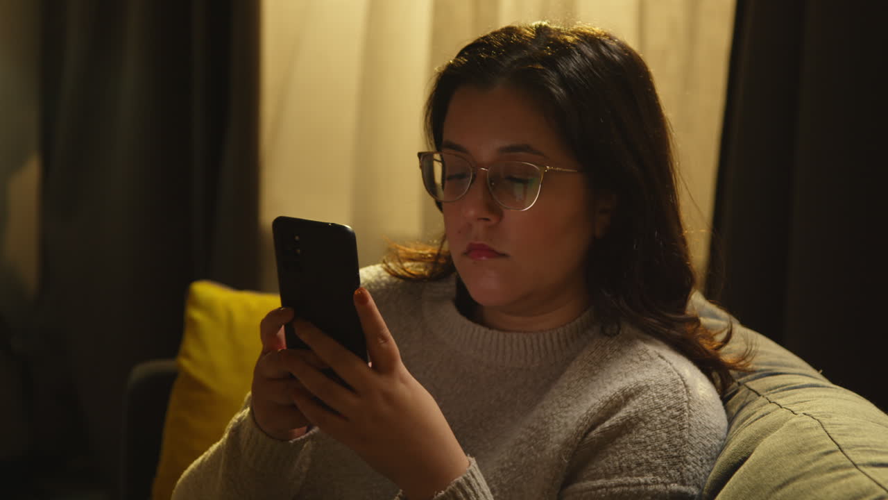 mujer joven en casa sentada en el sofá con el teléfono móvil desplazándose por internet o redes sociales en la noche 3