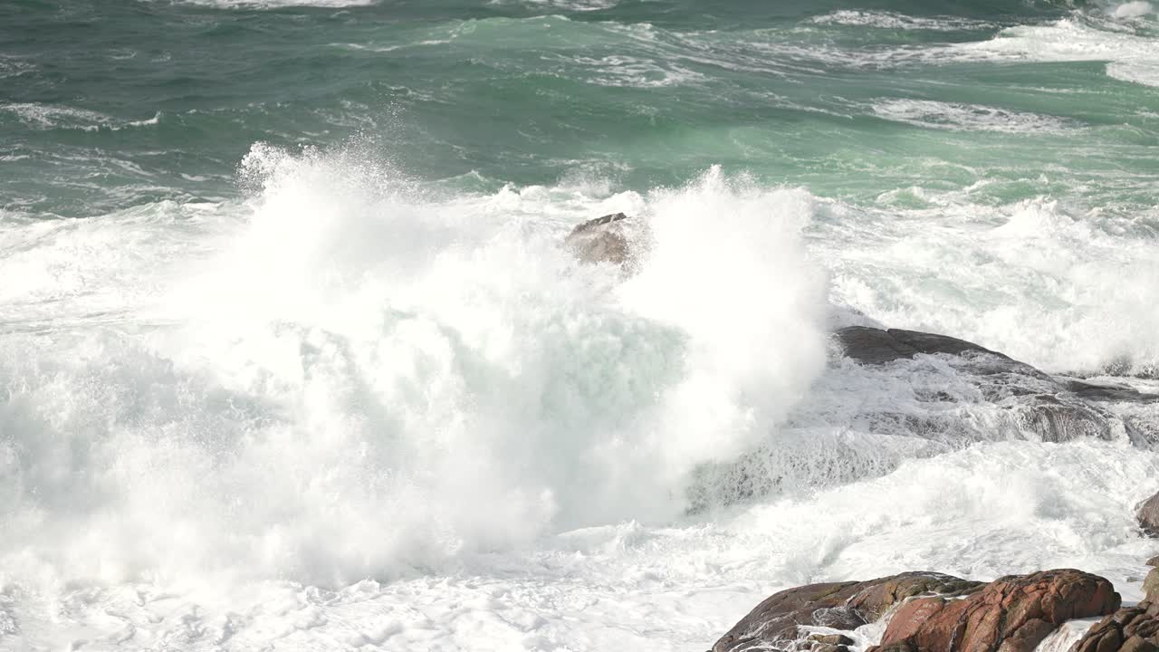 The scene shows a rocky formation surrounded by turbulent ocean waves on a stormy day. Water crashes against the rocks, creating a dramatic and wild atmosphere