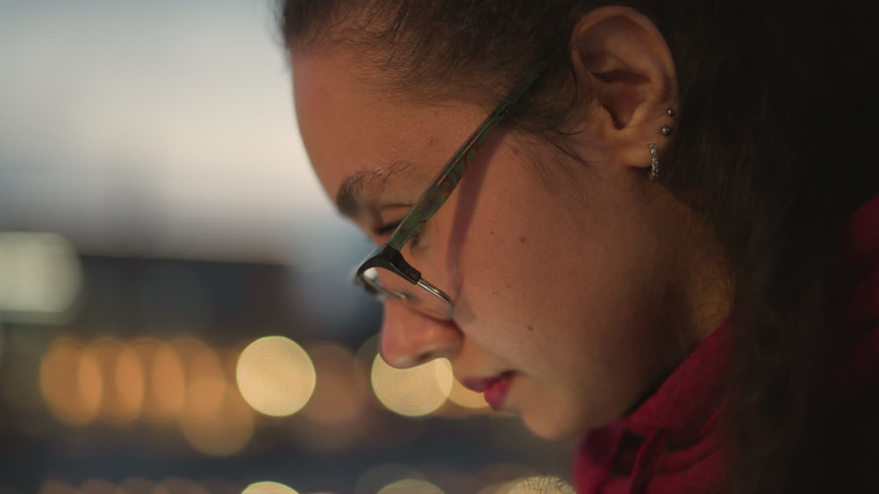 Young Woman Examining Screen At Night, Outdoor Scene Of Woman Concentrating On Device, Young Woman Intently Studying On Her Device During Nighttime With City Background And Reflective Glasses