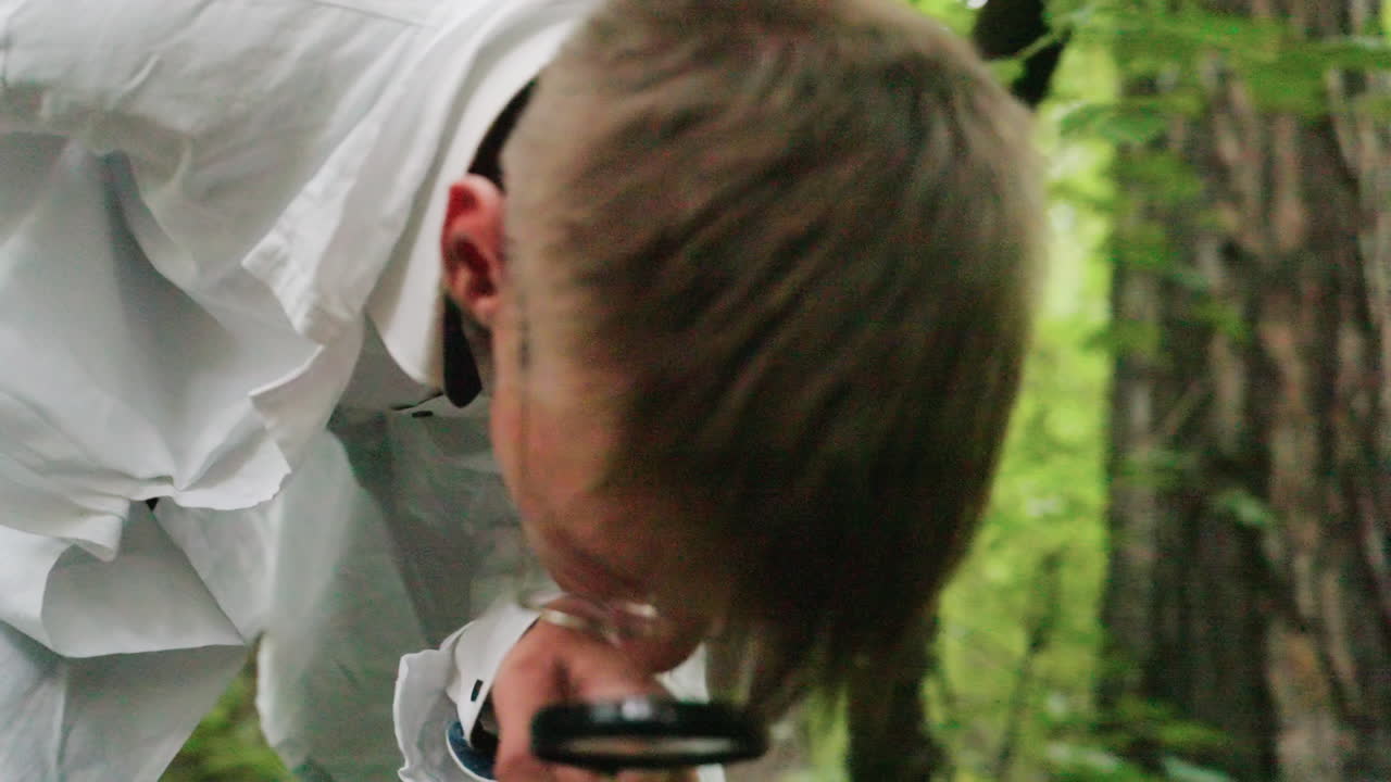 Scientific researcher in white coat and glasses holding microscope lens while observing closely in forest surrounded by green foliage, conducting detailed ecological study and outdoor environment