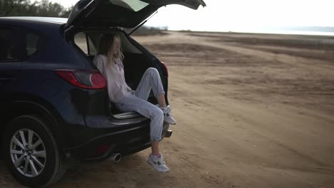 Woman-traveler-having-a-break-sitting-in-car-trunk,-looking-on-waterfront-on-horizon