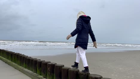 Frau-Hält-Das-Gleichgewicht-Auf-Einem-Holzpfahl-Am-Sandstrand-Neben-Tosenden-Wellen-Der-Nordsee-Bei-Grauem-Himmel-Und-Springt-Herunter