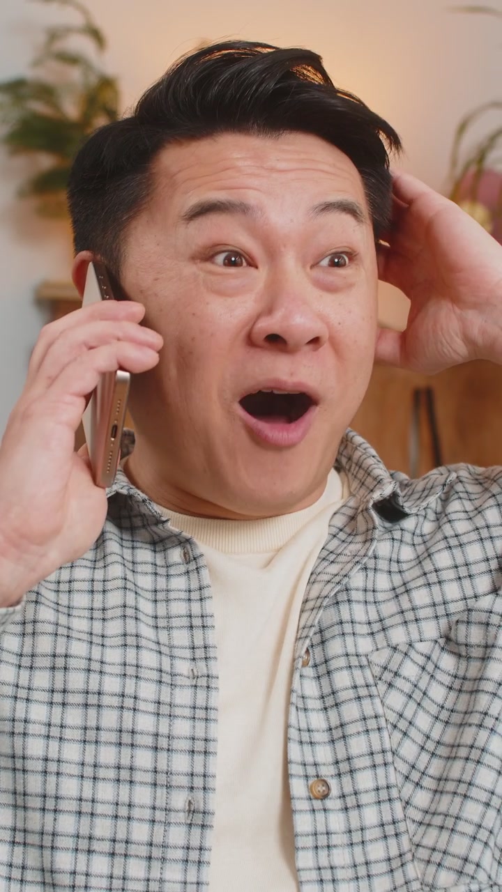 リビングのソファに座り、友人と電話で楽しく会話する笑顔の若い中国人男性。