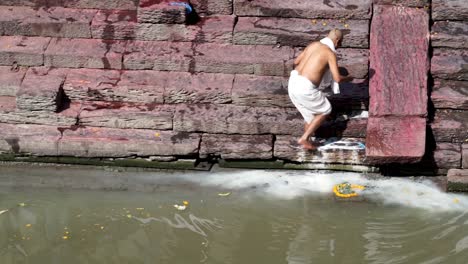 Old-Man-Washed-His-Dish-Pan-On-A-Shallow-River-Beneath-The-Old-Stone-Staircase-Of-Pashupatinath-Temple-In-Kathmandu,-Nepal