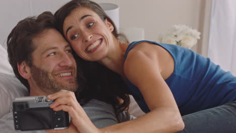 happy couple taking photos together using camera having fun at home on bed playfully enjoying romantic relationship photographing each other