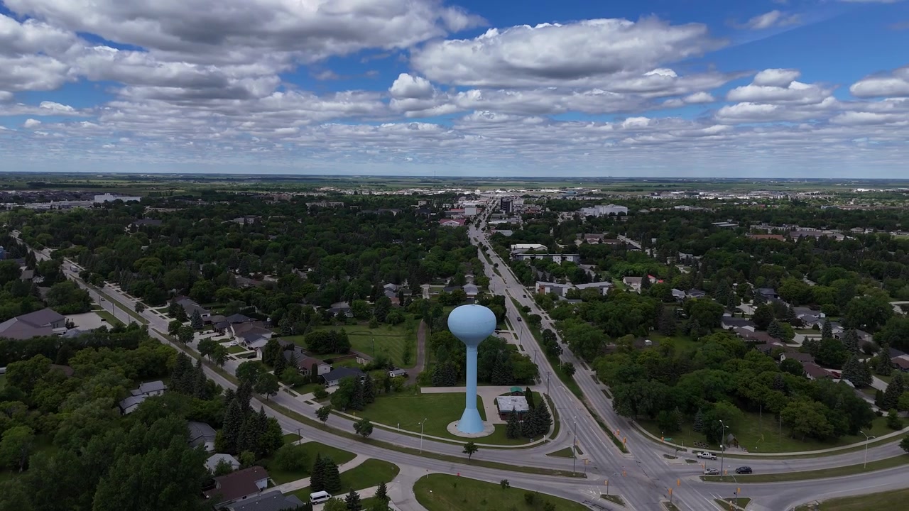 Drone roterer til venstre på afstand omkring det lyseblå vandtårn i Steinbach, Manitoba, der viser den hævede tank, veje, biler, huse, træer, forstæder og grønt græs om sommeren