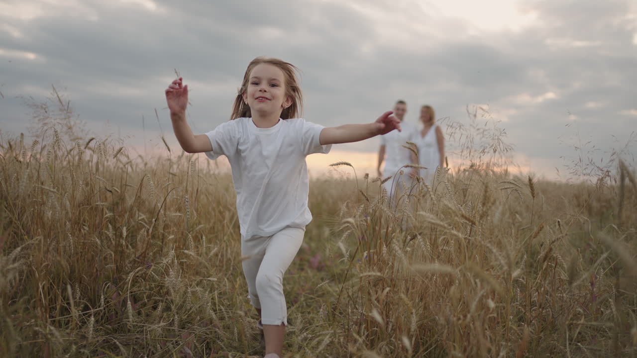 счастливая улыбающаяся девочка, смотрящая в камеру, бежит в замедленном движении на закате в поле зерновых ушей.