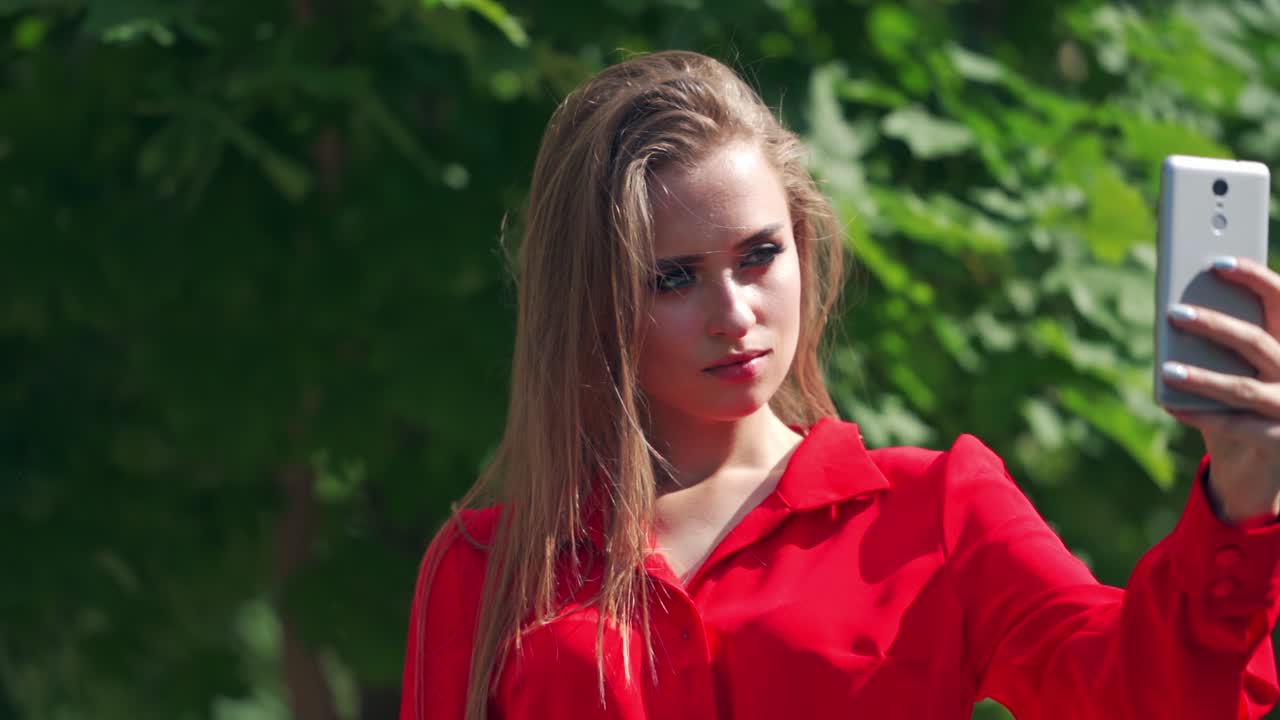Beautiful girl doing selfie outdoors. Attractive female holding mobile phone in her hand and photographing herself near the green tree. Close-up.