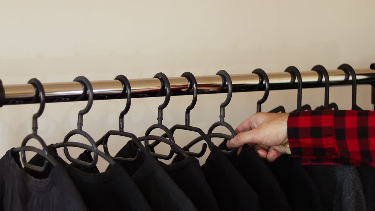 A man's hand, wearing a red plaid flannel shirt picks a black t-shirt from a rack full of black t-shirts