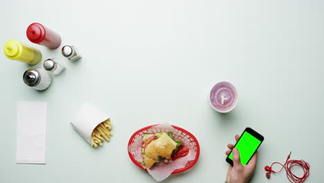 Vista-Superior-Hombre-Comiendo-Hamburguesa-Usando-Un-Teléfono-Inteligente-Con-Pantalla-Táctil-En-Un-Restaurante-De-Comida-Rápida-Americano-Manos-Desde-Arriba---Dragón-épico-Rojo